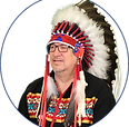 Chief Monias smiles under his feathered headdress banded in red. His shirt is black with a red yellow and teal beading across the chest