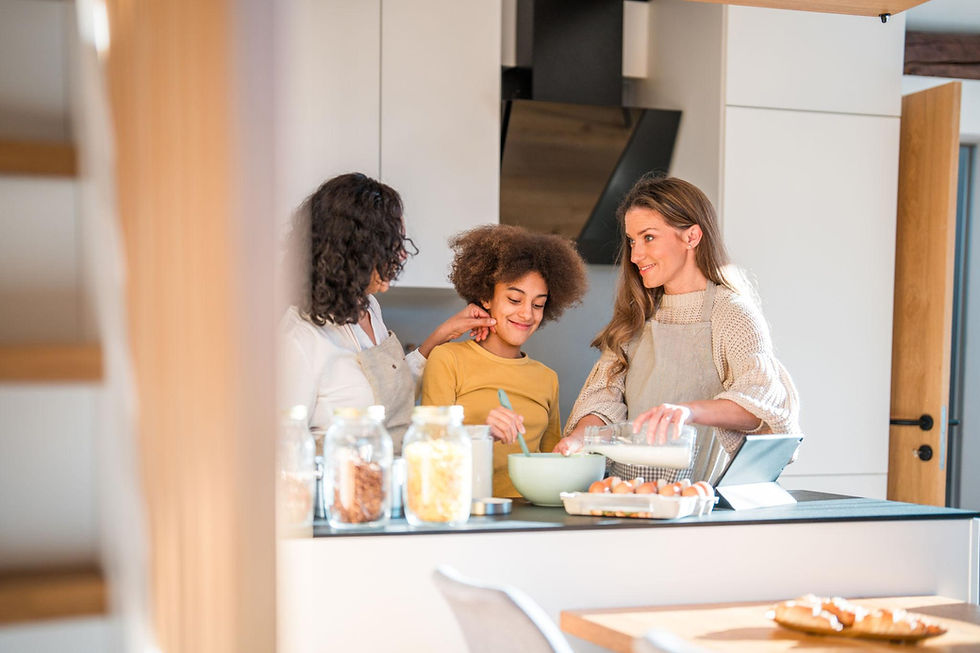 Three people cooking in a bright kitchen. One pours milk into a bowl while a child stirs. Jars and eggs are on the counter. Smiling.