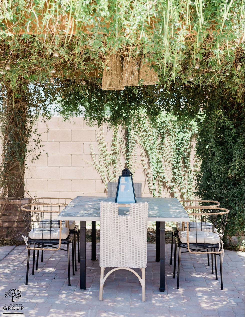 Outdoor dining area with a modern, rustic vibe featuring a rectangular table and a mix of woven and metal chairs. The space is sheltered by a lush, vine-covered pergola, creating a cozy and inviting atmosphere.