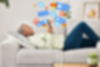 Woman lying on a couch looking at her phone and smiling with various cartoon bubbles showing alerts, emails, hearts, emojis for her digital content.