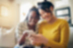 Two women sitting on a couch smiling and looking a cell phone.
