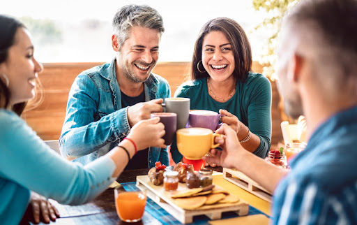 Group of four people, two men and two women, with smooth skin, doing a cheers with their coffee cups over a breakfast table.