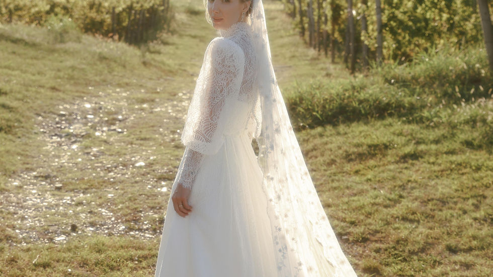 Bride in a white vintage lace gown and veil stands on a sunlit vineyard path, looking back. The setting is serene with green rows of grapevines.