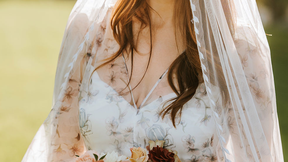 Bride in floral gown and veil holds bouquet of roses outdoors. Sunlit, serene expression, green field backdrop.