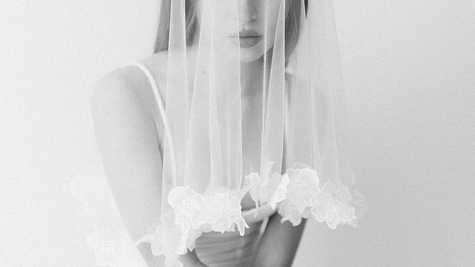 Bride in a white jumpsuit, wearing a floral veil, sits pensively against a soft gray background. The mood is serene and contemplative.
