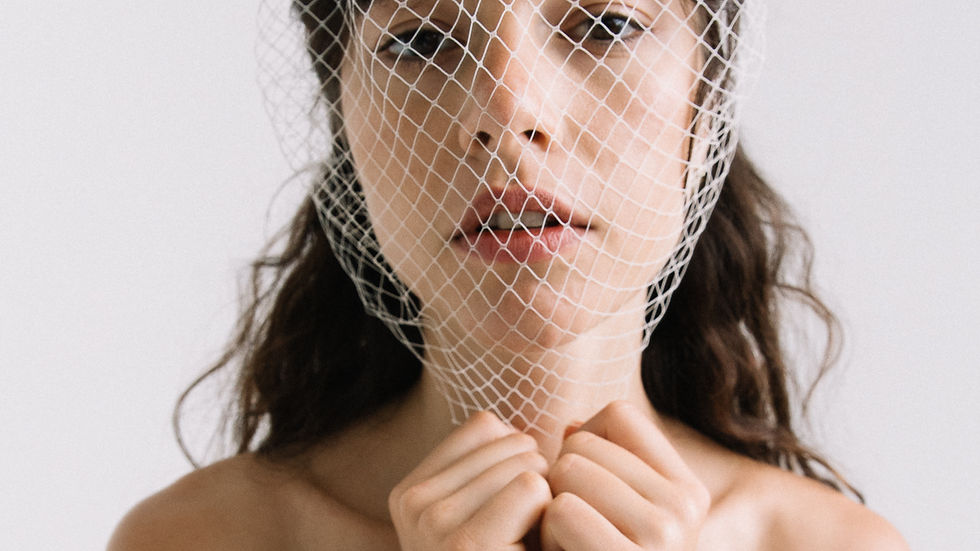 Bride with netted birdcage veil gazing in camera, holding veil at chin. Neutral expression, wearing white strapless dress. Minimalist light background.