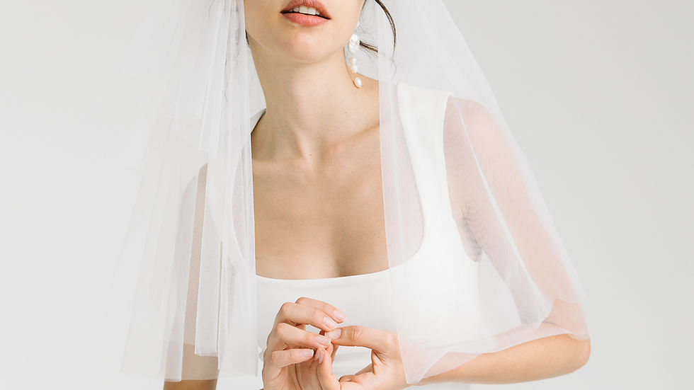 Bride in white tube dress and elbow length veil with bow, hands clasped, neutral expression. Simple white background, modern and minimal aesthetic