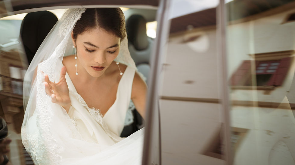 Bride in white dress and veil exiting a car, looking down. Italian architecture reflected in window. Elegant and serene mood.
