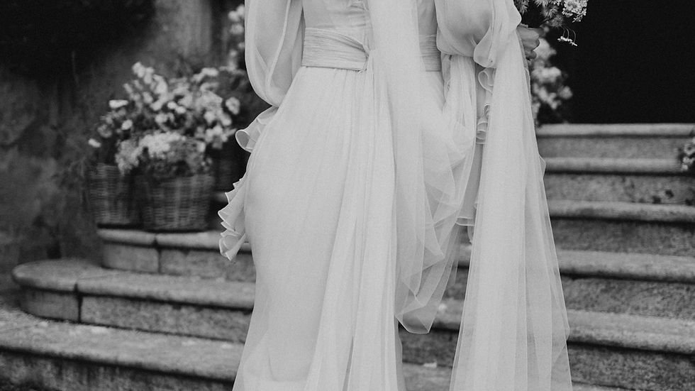 Bride in flowing gown with veil, holding a bouquet, walks up stone steps. Brick wall and flowers in background. Black and white photo.