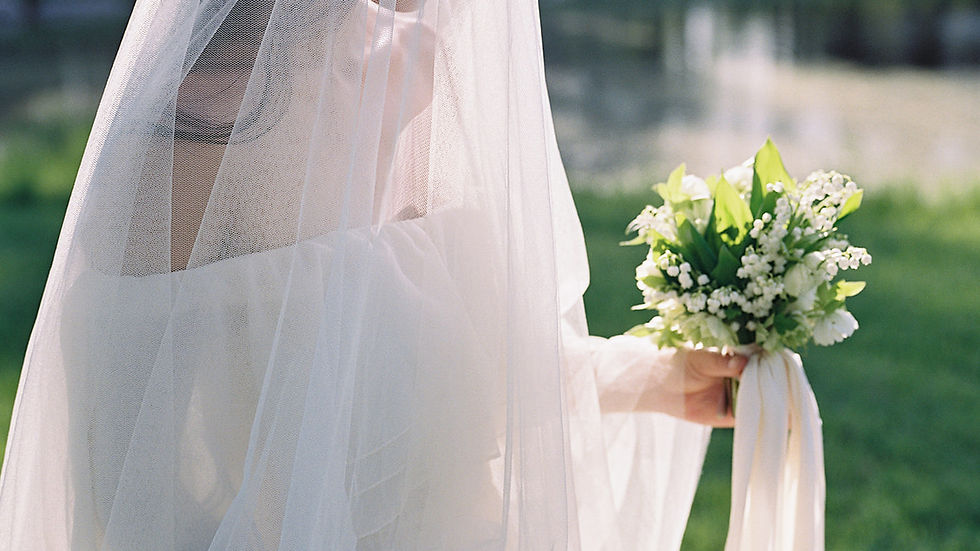 Bride in white veil holding a bouquet with lilies. Sunlit setting with a pond and grass in the background. Serene and elegant mood.