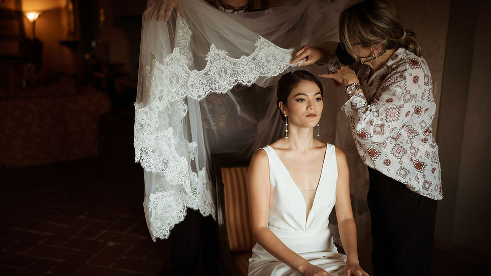 Bride in a white dress sits calmly as two people adjust a lace veil over her. Dimly lit room with an elegant, serene atmosphere.