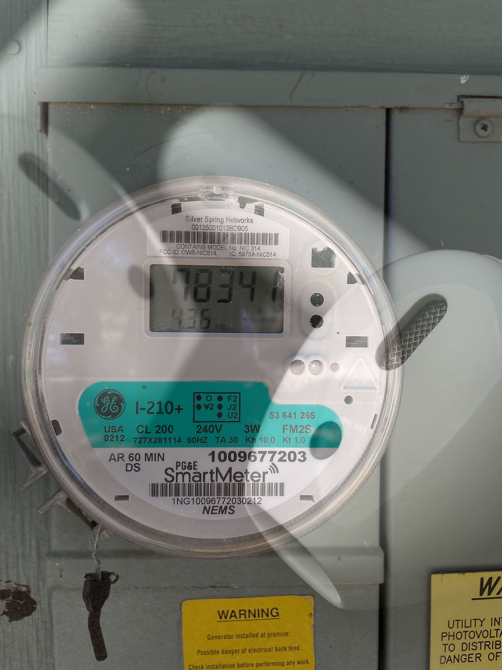 Close-up of a digital GE SmartMeter showing 78341 kWh. Labels and warning stickers visible. Mounted on a light gray panel. There is an overlay of ear buds one the image.