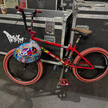 Dave Hill's red BMX bike with red tires and a heavily sticker-covered blue helmet on the seat, leaning against black road cases backstage, with a Puscifer graffiti logo painted on the case behind it.