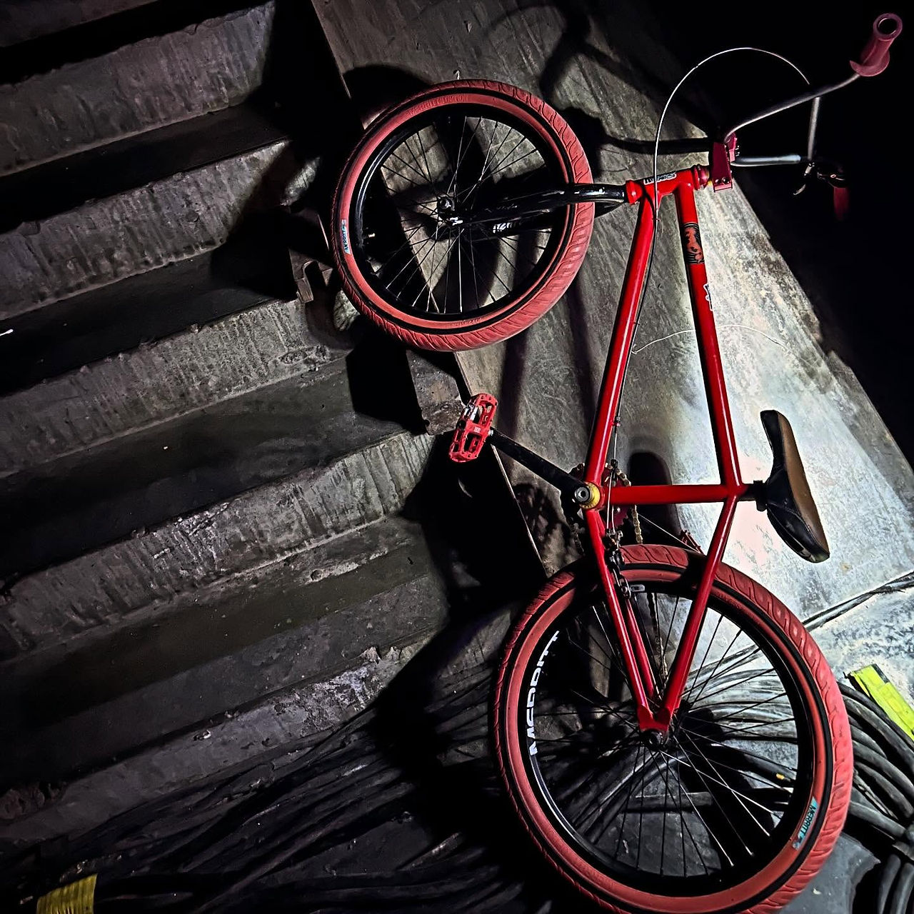 moody low-light shot of Dave Hill's red BMX bike with red tires leaning against a dark concrete stairwell, coiled cables on the floor beside it.