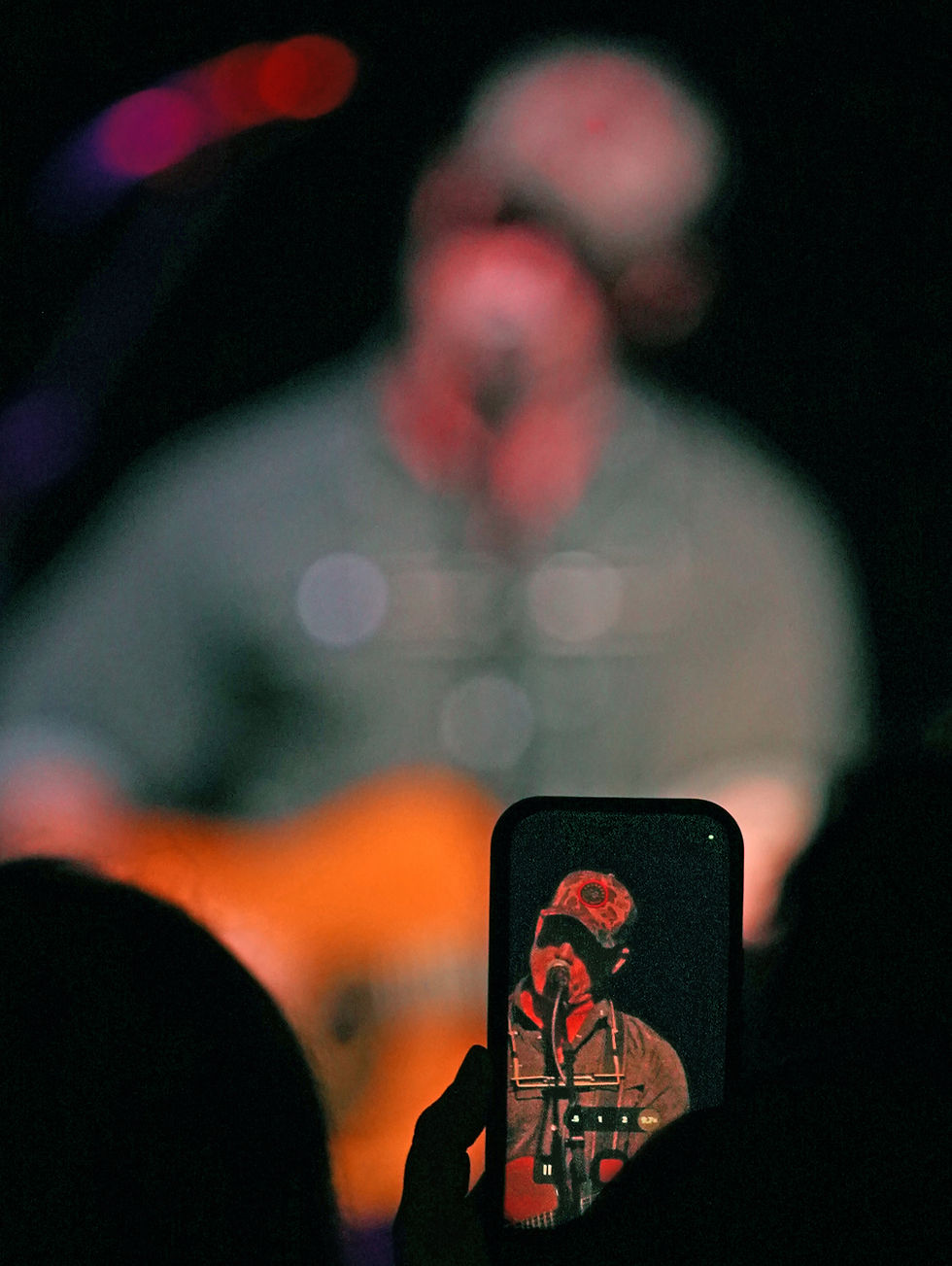 Audience member films a blurred singer on stage; phone screen shows the performer under red lights at a dark concert.