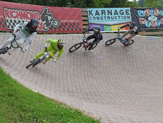 Four BMX racers lean through a banked turn on a paved track, with Richmond BMX and sponsor banners behind.