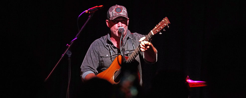 Man in a camo cap sings into a mic while playing acoustic guitar on a dark stage lit red and purple.