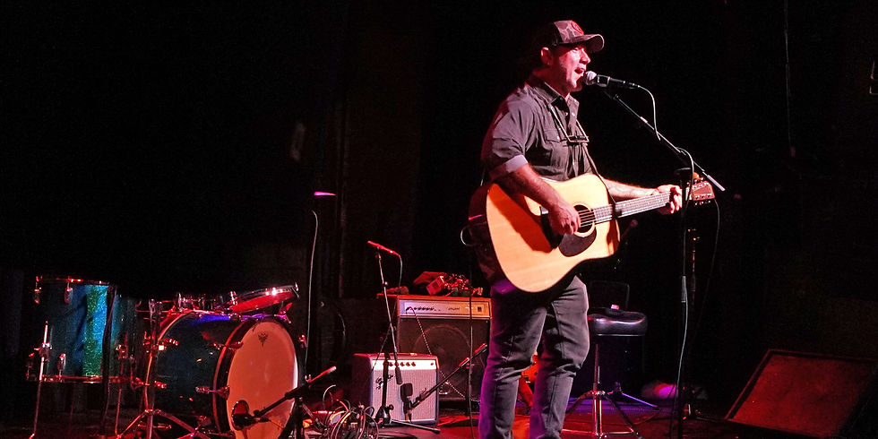 Man singing and playing acoustic guitar on a dark stage with drums and amps, lit by red and pink concert lights.