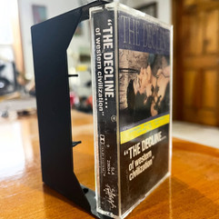 The Decline cassette case on a wooden table, with bold title and punk band names; cover shows a woman singing into a microphone.