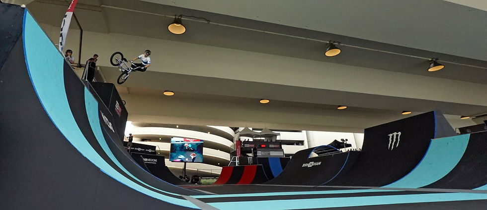 BMX rider midair over a blue-and-black ramp in an indoor skatepark, with spectators and Monster logos.