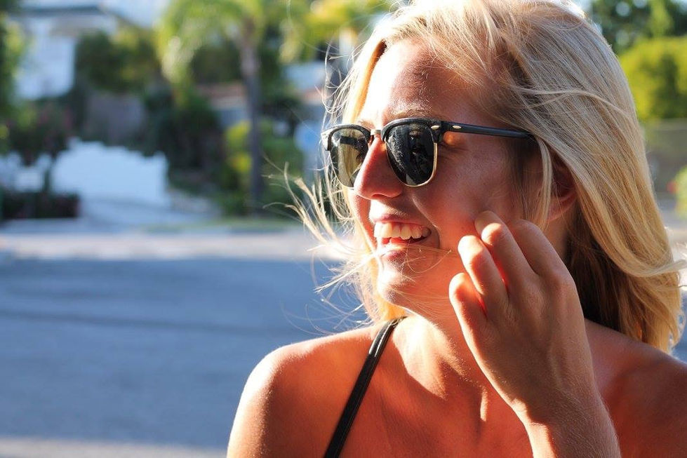 Close-up candid portrait of a laughing blonde woman wearing retro-style sunglasses, bathed in warm golden sunlight with a blurred suburban street and tropical trees in the background.