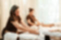 Two massage therapists performing leg and foot massages on clients in a serene spa setting. The therapists, dressed in brown uniforms, are focused on providing a relaxing experience, with soft natural lighting filtering through curtains in the background.