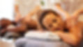 A woman with curly hair is lying on a massage table with her head resting on a folded towel, receiving a shoulder massage from a therapist. She appears relaxed with her eyes closed. In the background, a man is also lying on a massage table, receiving a back massage from another therapist. The setting is calm and spa-like, with a focus on relaxation and wellness.