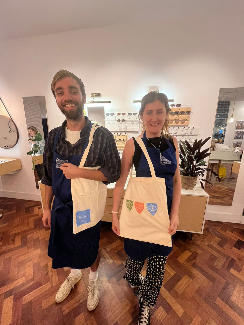 a man and a woman stand facing the camera, wearing tote bags that they have printed. 