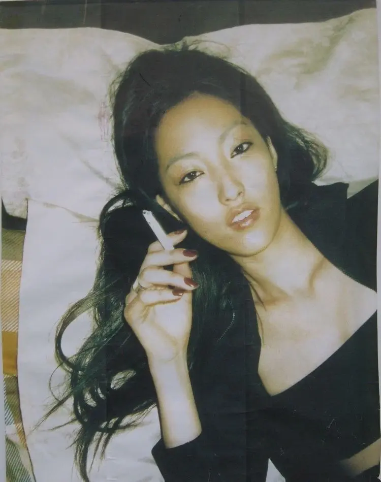 Retrato de una joven japonesa recostada sobre una cama, sosteniendo un cigarrillo y mirando a la cámara, en una fotografía de estética íntima asociada a la cultura visual japonesa de los años noventa.