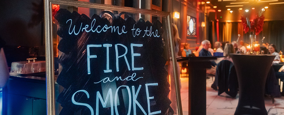 Welcome sign reading “Fire and Smoke” at an upscale event with high-top tables and guests in the background.