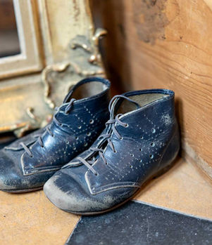 Abgenutzte dunkelblaue Kinderschuhe vor einem alten goldenen Spiegelrahmen auf gemustertem Boden.