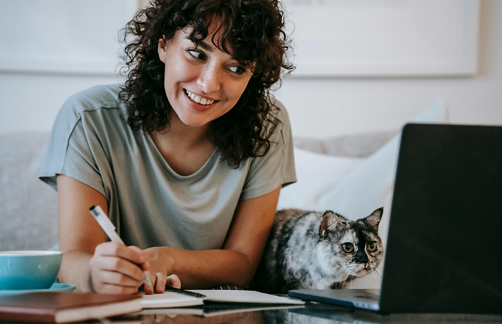 Mulher sorridente trabalhando em um laptop enquanto faz anotações, com um gato ao lado, em um ambiente caseiro