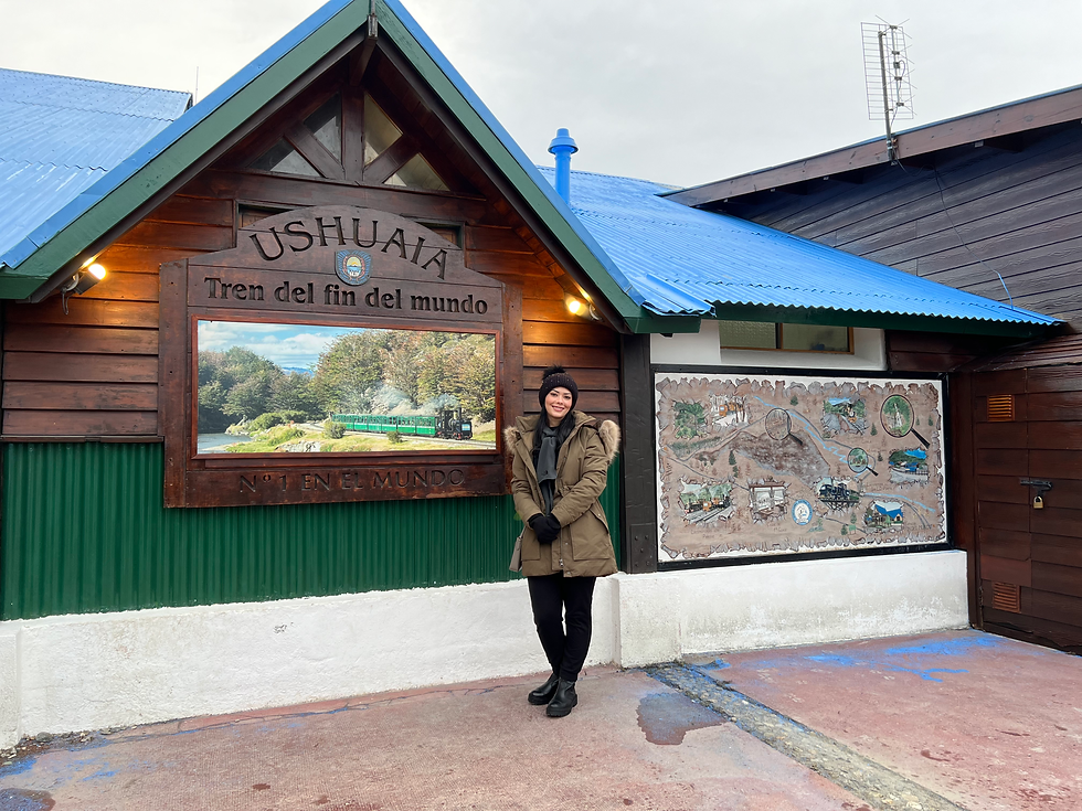 Mulher sorridente vestindo roupas de frio posa em frente à estação do “Tren del Fin del Mundo”, em Ushuaia. Ao fundo, uma placa de madeira com a inscrição “Ushuaia - Tren del Fin del Mundo - N°1 en el mundo” e uma imagem do trem verde rodeado pela natureza.