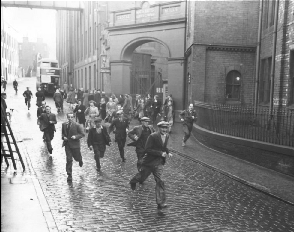 Black-and-white photo of workers leaving the North British Rubber Company’s Castle Mills factory in Edinburgh during the 1950s. Men in flat caps and suits walk and run along a cobbled street outside the factory gates, with a vintage double-decker bus in the background.