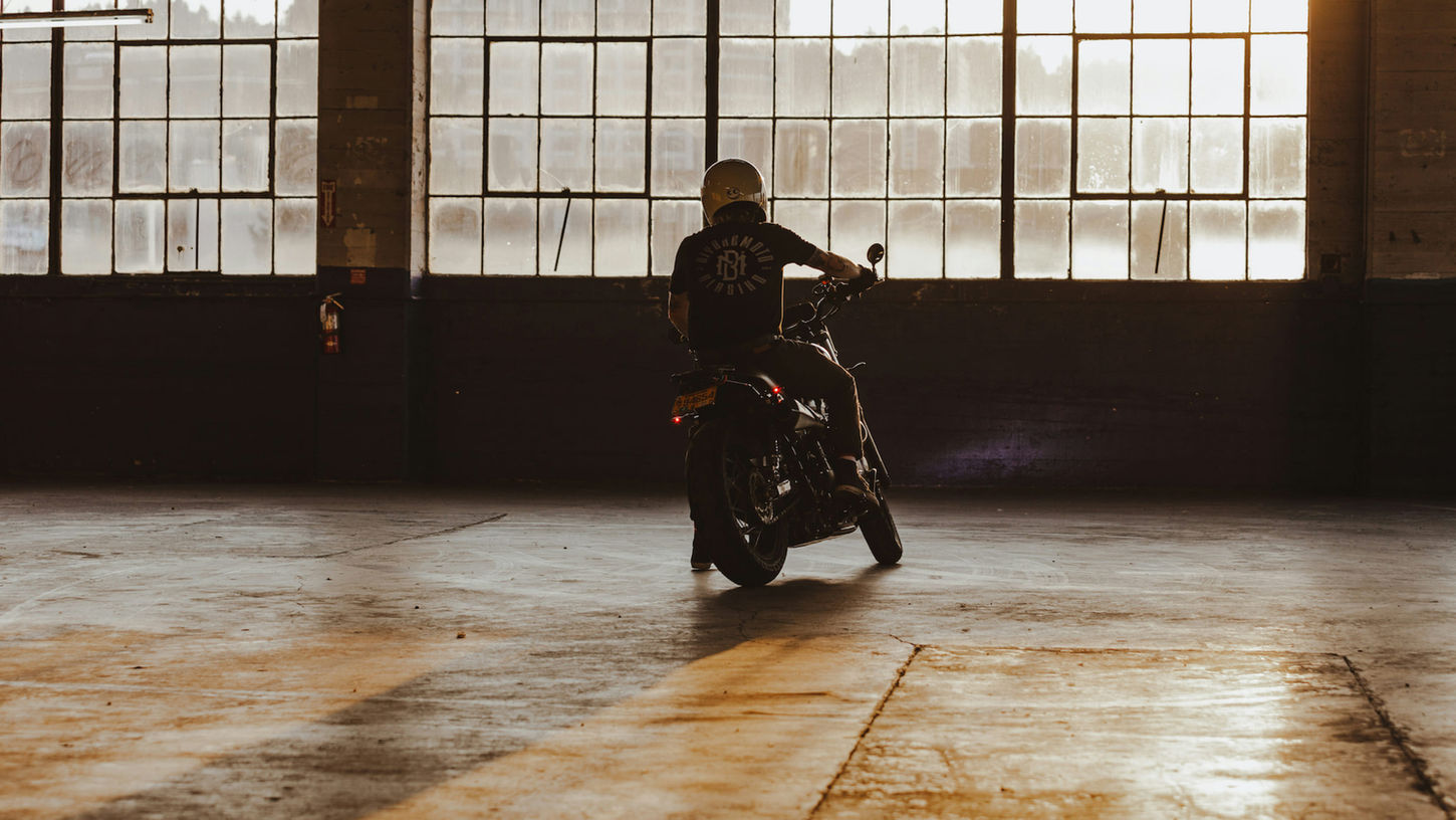 Motorcyclist rides inside spacious warehouse. Sunlight filters through large windows, casting long shadows for a warm, moody ambiance.