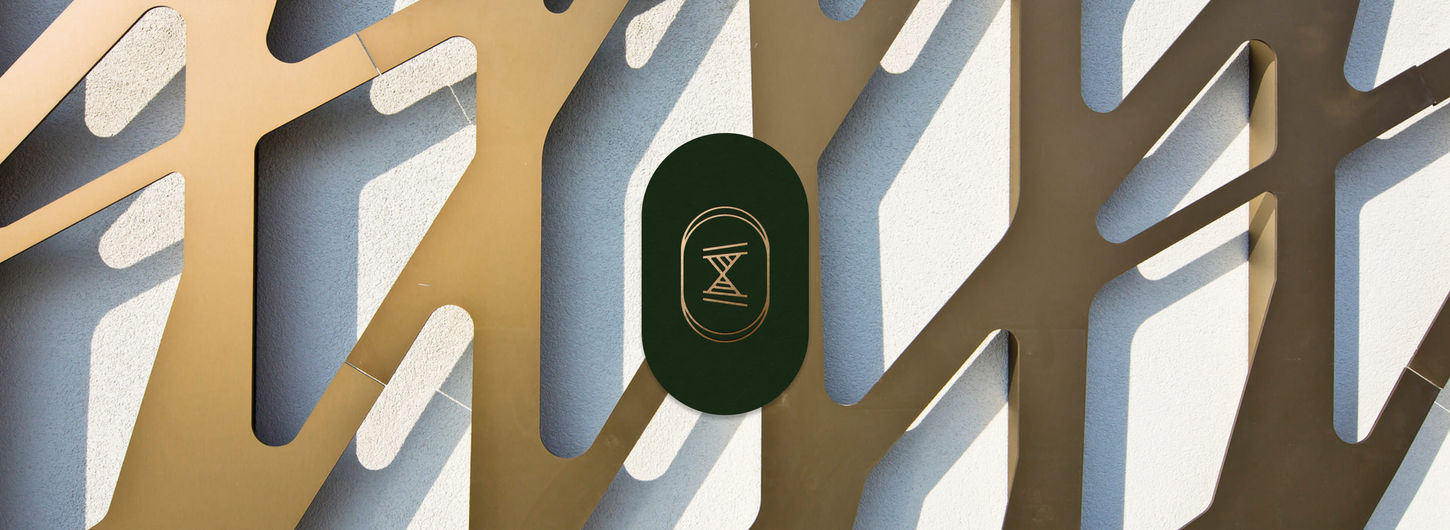 Abstract design with golden, angular cutouts on a textured white wall. A green oval with a geometric Hora Residences symbol creates contrast, evoking modernity.