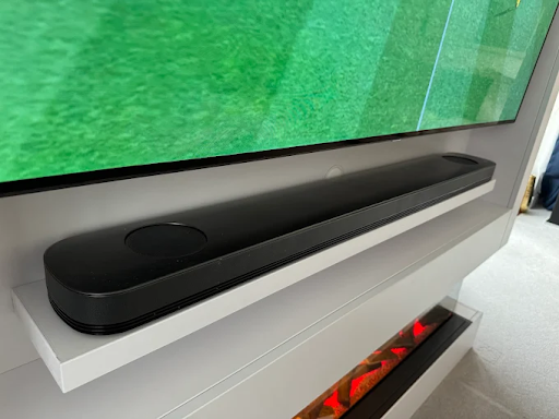 Black soundbar on white shelf below a large TV showing green content. Below, a lit electric fireplace adds a warm touch.