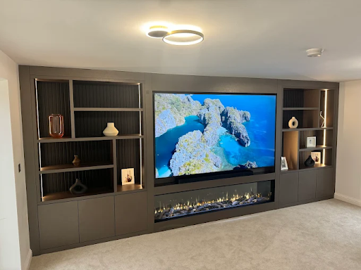 Modern living room with large TV displaying an ocean view. Built-in shelves hold vases and photos. A sleek electric fireplace adds ambiance.
