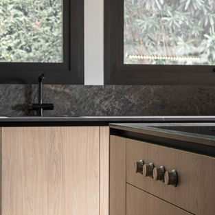 cuisine au design contemporain qui privilégie un équilibre raffiné entre les textures naturelles et industrielles. L’aménagement s’articule autour de l’utilisation de matériaux contrastés, mariant la chaleur organique du bois clair à la rigueur minérale de plans de travail en pierre sombre. La structure privilégie une ouverture sur l'extérieur, intégrant de larges fenêtres qui encadrent la végétation comme un élément décoratif à part entière. À travers ces différentes perspectives, l'œuvre souligne une esthétique de minimalisme sophistiqué où chaque détail fonctionnel s'efface derrière une harmonie visuelle apaisante.