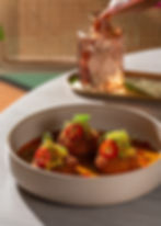 A bowl of golden croquettes topped with sliced red chilies and greens, in a rich orange sauce. In the background, a glass decanter.