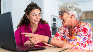 Two women smiling at a laptop; one in a pink shirt, the other in floral. The older woman points at the screen. Bright room setting.