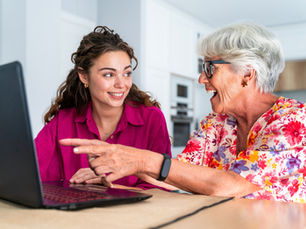 Two women smiling at a laptop; one in a pink shirt, the other in floral. The older woman points at the screen. Bright room setting.