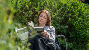 Woman reading a book in a chair amid lush green bushes, looking calm and relaxed outdoors.