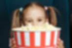 Close up of a young child with pigtails holding a large red and white striped container of popcorn in front of her face