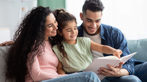 A smiling family sits on a sofa reading a book together. The child points at a page. Cozy living room setting with soft lighting.