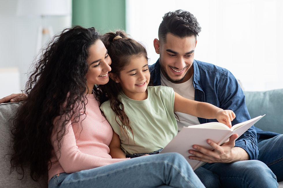 A smiling family sits on a sofa reading a book together. The child points at a page. Cozy living room setting with soft lighting.