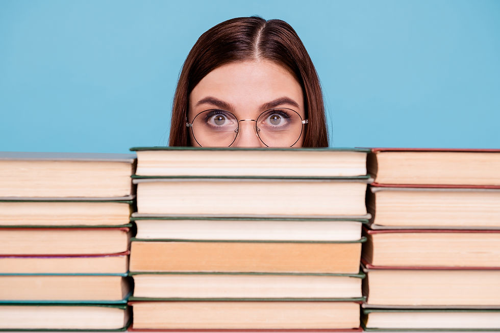 Woman with glasses peers over a stack of books against a blue background, conveying curiosity and focus.