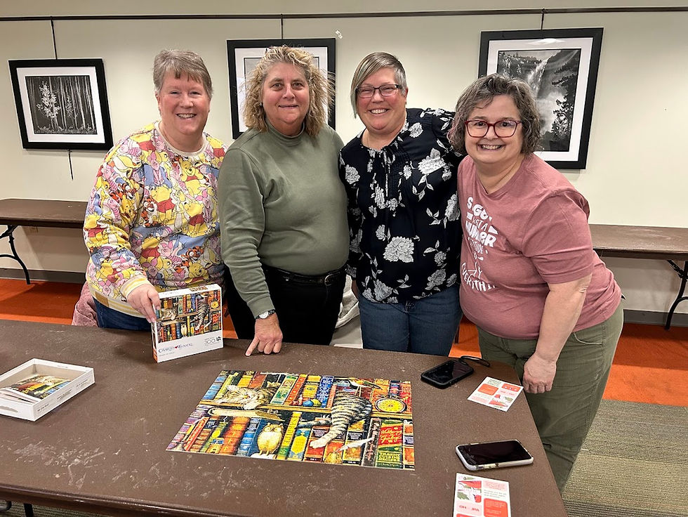 Four smiling women stand around a completed colorful puzzle on a table. The setting is indoors with framed photos on the wall.