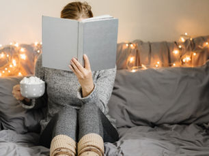 Person in cozy sweater reads a book on a bed with fairy lights, holding a mug of marshmallows. Relaxed, warm ambiance.