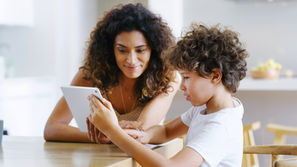 Mother and young boy in a bright kitchen look at a tablet together, smiling softly.